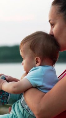 Anne, yaz akşamı nehrin kenarında oturan bir bebeği kollarında tutuyor. Annesinin oğlunu kafasından öpmesi. Arkaplan bulanık. Dikey video