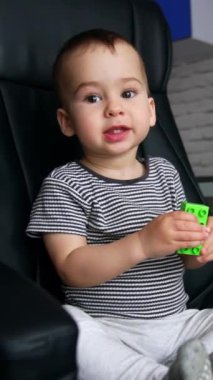 Büyük bir ofis koltuğunda oturan mutlu gülümseyen sevimli çocuk. Sevimli bebek sandalyeyi açıyor ve bir oyuncakla oynuyor. Dikey video