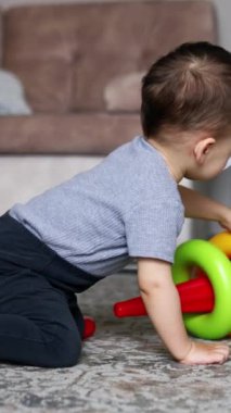 Bebek halının üzerinde parlak oyuncak piramidiyle oynuyor. Çocuk oyuncağı parçalara ayırıyor ve tekrar inşa ediyor. Kara kedi yanında oturuyor. Dikey video
