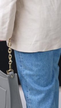 Altın zincir saplı, güzel, gri deri bir çanta. Ceketli ve kot pantolonlu bir kadın elinde modern bir çanta tutuyor. Dikey video