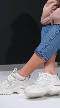 Siyah basamakta oturan bir kızın kırpılmış görüntüsü. Stüdyoda kot pantolon, sweatshirt ve beyaz spor ayakkabı mankeni. Dikey video