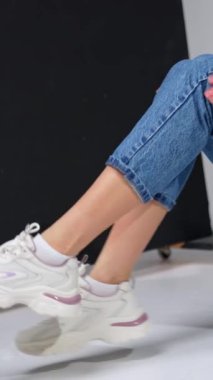 Tarz sahibi, sıradan görünümlü, tanınmamış bir kadın model. Kız kot pantolon, pembe kazak ve beyaz spor ayakkabı giyiyor. Stüdyo çekimi. Dikey video