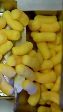 Taşıyıcı bantta lezzetli mısır çubukları. Otomatikleştirilmiş gıda fabrikasında içi sarı atıştırmalıklarla dolu metal kutular. Dikey video