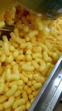 Bir sürü sarı gevrek mısır çubuğu taşıma hattına düşer. Fabrika işçisi aperatifleri kemer boyunca itiyor. Yiyecek fabrikası üretim konsepti. Dikey video