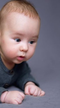 Gri elbiseli tatlı beyaz çocuk yatağın üzerinde yatıyor. Güzel çocuk şaşkınlıkla etrafına bakıyor. Gri zemin. Dikey video