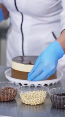 Şekerci tarafından süslenen beyaz ve çikolatalı pasta. Eldivenli kadın plastik bardaklardan farklı şekerler alıyor ve onları pastanın üstüne serpiyor. Dikey video