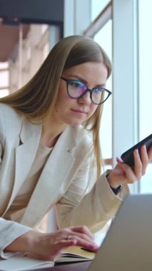 Gözlüklü uzun saçlı kadın hemen telefon ve dizüstü bilgisayar kullanıyor. Bayan masanın üstünde duruyor ve bilgisayarda bir şeyler yazıyor. Dikey video