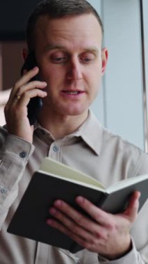 Gri gömlekli adam telefon görüşmesi yapıyor. Erkek çalışan önünde kağıt bir defter tutuyor ve telefonda konuşuyor. Dikey video