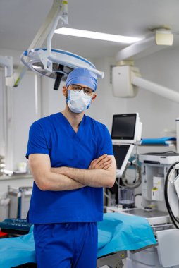 Cerrah ameliyathanede duruyor. Ameliyat odasında önlüklü ve maskeli bir erkek cerrah kollarını kavuşturup duruyor..