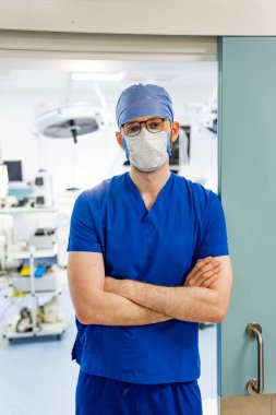 Cerrah, ameliyathane kapısında bekliyor. Ameliyat önlüğü takan, ameliyat şapkası takan ve yüz maskesi takan bir cerrah kollarını bir ameliyathanenin kapısında kavuşturur..