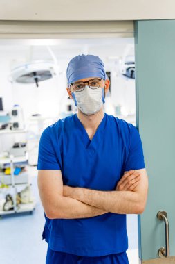 Cerrah ameliyathanede bekliyor. Ameliyat odasında kolları kavuşturulmuş önlüklü ve maskeli bir cerrah..