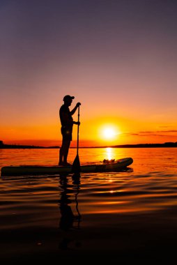 Sunset 'te sörf tahtası. Gün batımında gölün ortasında ayakta duran bir raket. Güneş onların ardından batıyor ve suyu yansıtıyor..