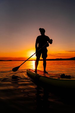 Gün batımında Paddleboder 'ın silueti. Bir insan, gün batımına karşı siluet içinde bir kürek tahtasında duruyor, sakin suda kürek çekiyor..
