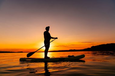 Sunset 'te sörf tahtası. Bir adam durgun bir gölün üzerinde, canlı bir günbatımına karşı duran bir kürek tahtasında duruyor..