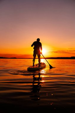 Sunset 'te sörf tahtası. Güneş batarken bir adam gölün üzerinde kürek çekiyor ve suyun üzerine sıcak bir parıltı saçıyor..