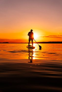 Sunset 'te sörf tahtası. Durgun bir gölün üzerinde tek bir kişi güneş batarken suda güzel bir turuncu ışıltı yaratıyor..
