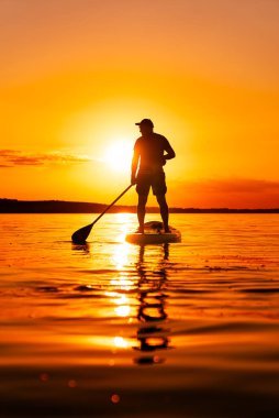 Sunset 'te sörf tahtası. Yalnız bir adam, turuncu bir günbatımına karşı silueti olan bir kürek tahtasının üzerinde duruyor. Su gökyüzünün canlı renklerini yansıtır..