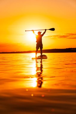 Sunset 'te sörf tahtası. Güneş arkalarında batarken sakin bir gölde kürek çeken bir insan, suyun üzerinde sıcak bir parıltı saçar..