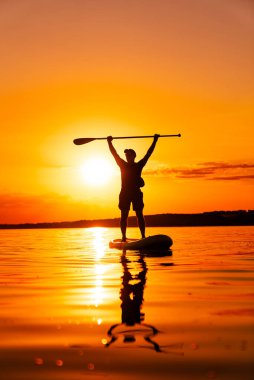 Sunset 'te sörf tahtası silueti. Bir adam, kolları havada, sakin bir gölün üzerinde canlı bir günbatımına karşı duran bir kürek tahtasında duruyor..