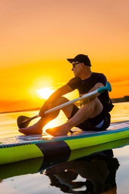 Sunset 'te sörf tahtasında dinlenen adam. Bir adam sakin bir gölde, günbatımının tadını çıkarıyor..