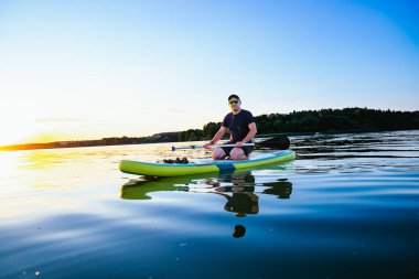 Man Paddleboard, Sunset Gölü 'nde. Bir adam gün batımında sakin bir gölün üzerinde ayakta duran bir kürek çeker. Su altın ışığı yansıtır..