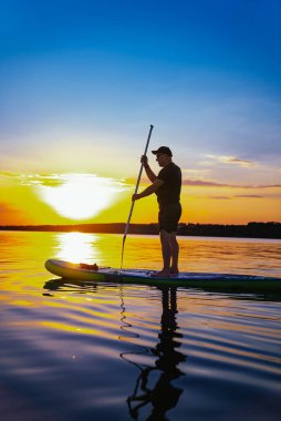 Sunset 'te sörf tahtası. Gün batımında sakin bir gölde elinde kürekle kürek çeken bir adam..