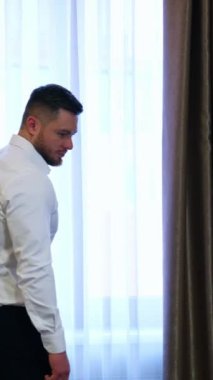 Pencereye yakın başarılı bir iş adamı. Modern bir otelde beyaz gömlekli yakışıklı bir adam. Lüks perdelerin arkasındaki sakallı genç adam. Dikey video
