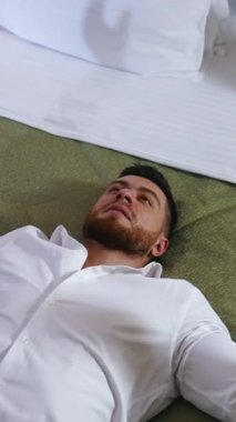 Genç iş adamı yatakta dinleniyor. Beyaz gömlekli yakışıklı adam bir otel odasında tek başına dinleniyor. İş gezisi sırasında tatil. Dikey video