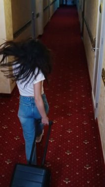 Bavullu mutlu kadın. Neşeli genç bayan gezgin elinde bavulla otel koridorunda yürüyor ve neşeyle zıplıyor. Üst Manzara. Dikey video
