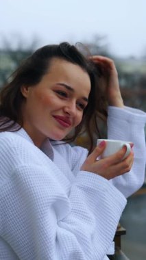 Otelde kaliteli vakit geçiren neşeli bir kadın. Balkonda bornozlu genç bir kadın sabahları arka planda kahve içiyor. Yan görüş. Dikey video