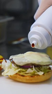 Fast food restoranında hamburger yapmak. Hamburgerin üzerine sos sürüp, üstünü kızarmış ekmekle kaplıyor. Yakın plan. Lezzetli hamburgerler yemeğe hazır. Dikey video