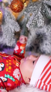Kırmızı elbiseli ve çizgili şapkalı sevimli bir erkek bebek sırt üstü yatıyor. Sağlıklı çocuk süslü Noel ağacının tadını çıkarıyor. Dikey video