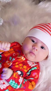 Kırmızı beyaz şapkalı, ciddi bir erkek bebek bembeyaz ekose kumaşın üzerinde yatıyor. Süslü Noel ağacının yanındaki komik çocuk. Yukarıdan bak. Dikey video