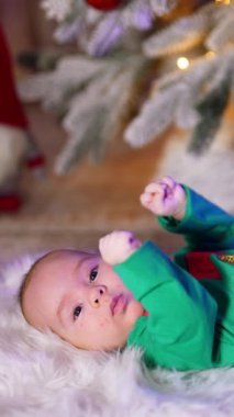 Yeşil kostümlü sevimli çocuk ekose üzerinde huzur içinde yatıyor. Arkaplanda bulanık bir şekilde süslenmiş Noel ağacı. Dikey video
