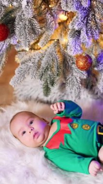 Yeşil elf kostümü içindeki beyaz çocuk ve tüylü ekose üzerinde geyik dinlenen kırmızı çoraplar. Güzel bebek Noel ağacının altında etkin bir şekilde bacaklarını hareket ettiriyor. Dikey video