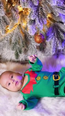 Elf yeşili kostüm, köknar ağacının altında yatan bir çocuğu giydirdi. Arka planda süslü Noel ağacı ve oyuncak cüceler. Kara kedi geliyor. Dikey video