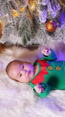 Yeşil elf kostümü giydirilmiş sevimli küçük bir çocuk. Sevimli çocuk Noel ağacının altında uzuvlarını hareket ettiriyor. Dikey video