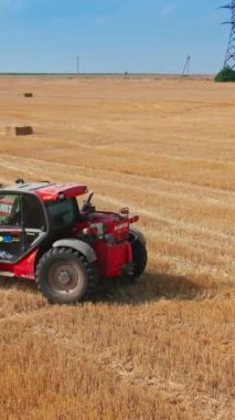 Yüksek güçlü patinaj yükleyicisi aynı anda iki ağır saman balyasını kaldırıyor. Araç, saman paketlerini traktöre taşıyor. Yeşil orman arka planı. Dikey video