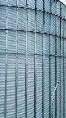 Modern silo fabrikasının bölgesinde duran dev bir gümüş metal ambar tankı boyunca yükseliyor. Çiftlik arazilerinin arka planındaki asansörlerin eğimli çatılarına bak. Dikey video
