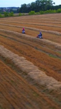 Erkek ve kız hasat döneminden sonra buğday tarlasında motosiklet sürüyor. Dişi binici arkadaşının önüne geçiyor. Köy ve ağaçlar arka planda. Dikey video