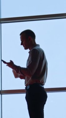 Adam masada oturuyor, telefonuna bakıyor, konuşuyor ve odada birine gülümsüyor. Düşük açı. Arka planda panoramik pencereler. Dikey video