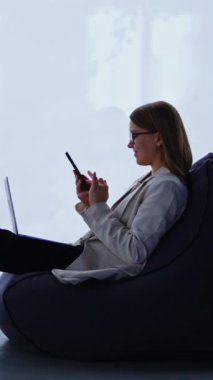 Ceketli ve kot pantolonlu bir kadın laptopuyla işten uzaklaştırılmış. Genç bir kadın telefonunda bir şey kaydırıyor. Panoramik pencere arkaplanı. Dikey video