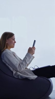 Genç bir kadın koltuğa rahatça oturur ve bacaklarını mindere uzatır. Bayan telefonunu tutuyor ve gülümsüyor. Dizüstü bilgisayar ve gözlükler dizlerinde. Arka planda panoramik pencereler. Dikey video