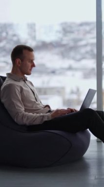 Orta yaşlı erkek, laptopta fasulye koltukta oturuyor. Adam dikkatini dağıtır ve yana bakar, elini alnına koyar. Arka planda bulanık şehir manzarası. Dikey video