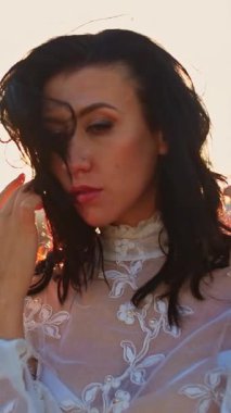 Beyaz elbiseli üzgün, düşünceli bayan, koyu saçlarına nazikçe dokunuyor. Parlak batan güneşe karşı dağlarda poz veren bir kadın. Kapatın. Dikey video