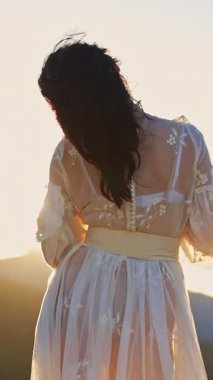Dağın tepesinde duran saydam beyaz elbiseli, siyah saçlı kadın. Tanımlanamayan bayan kameraya arkasını döner ve güneşin batışının tadını çıkarır. Dikey video