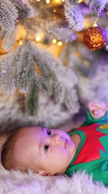 Elf yeşili kostüm giyen güzel bebek Noel ağacının altında yatıyor. Sakin çocuk süslemelere bakmış. Dikey video