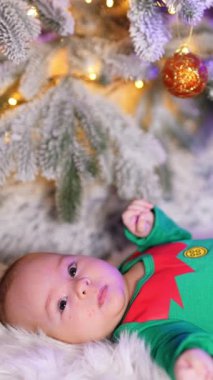 Yeşil elf takım elbiseli beyaz çocuk yeni yıl ağacının altında yatıyor. Noel süslemeleri içinde yeni doğmuş bir çocuk. Dikey video