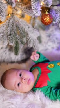 Yeşil elf kostümü içindeki küçük tatlı çocuk güzel dekore edilmiş Noel ağacının altında beyaz ekose üzerinde yatıyor. Sevimli çocuk ayaklarını sallıyor ve kameraya bakıyor. Dikey video