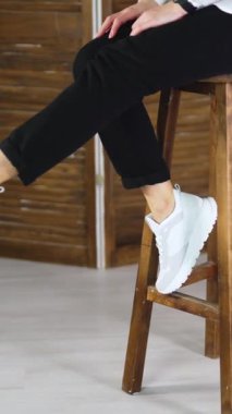 Kadın yüksek bir taburede oturuyor ve beyaz ayakkabılarıyla ayaklarını sallıyor. Model tabuetten atlıyor ve rahat bir ayakkabıyla uzaklaşıyor. Dikey video